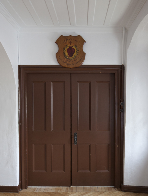 Porte brune double. Poignée en fer forgé. Au-dessus, une plaque de bois, dévotion au Sacré-Cœur en forme de bouclier.